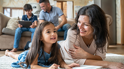 A mother and daughter are lying on their tummies on the floor reading a book together, while dad and their son are watching a digital screen in the background